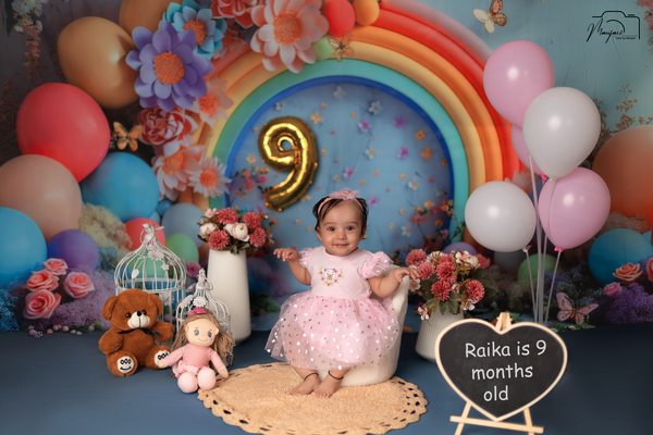 Baby seated beneath a rainbow balloon milestone display