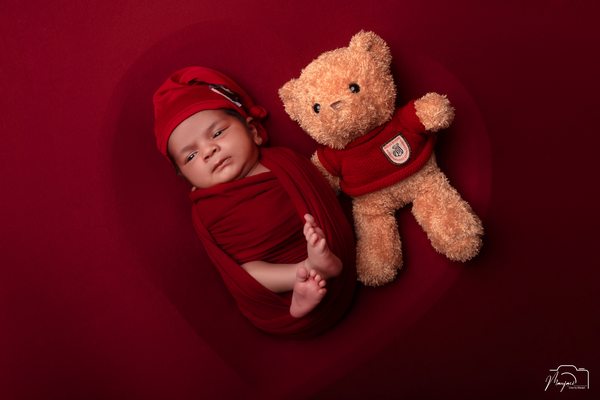Sleeping newborn wrapped in red beside a teddy bear